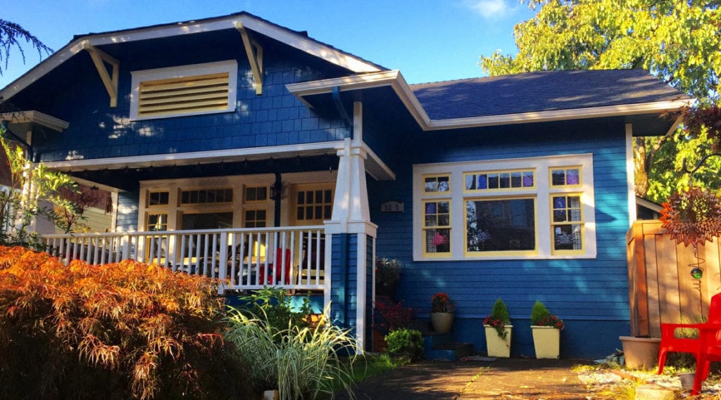 A charming blue Craftsman-style house with white trim, a covered front porch, and a small garden built by a skilled general contractor sits amid shrubs and plants in the yard on a sunny day.