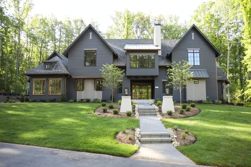 Large modern dark gray house with multiple gabled roofs, big windows, and a covered front porch, expertly crafted by a general contractor. A stone path winds through the neatly trimmed lawn and landscaped bushes to the entrance.