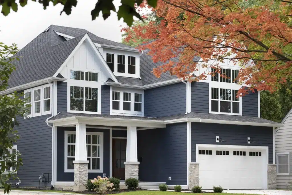 A modern two-story house with dark blue siding, white trim, and large windows built by a skilled general contractor. The white garage and front porch with stone-based columns complement the tree with orange leaves in the foreground.