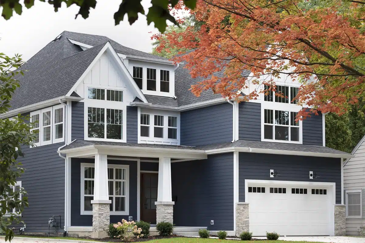 A modern two-story house with dark blue siding, white trim, and large windows built by a skilled general contractor. The white garage and front porch with stone-based columns complement the tree with orange leaves in the foreground.
