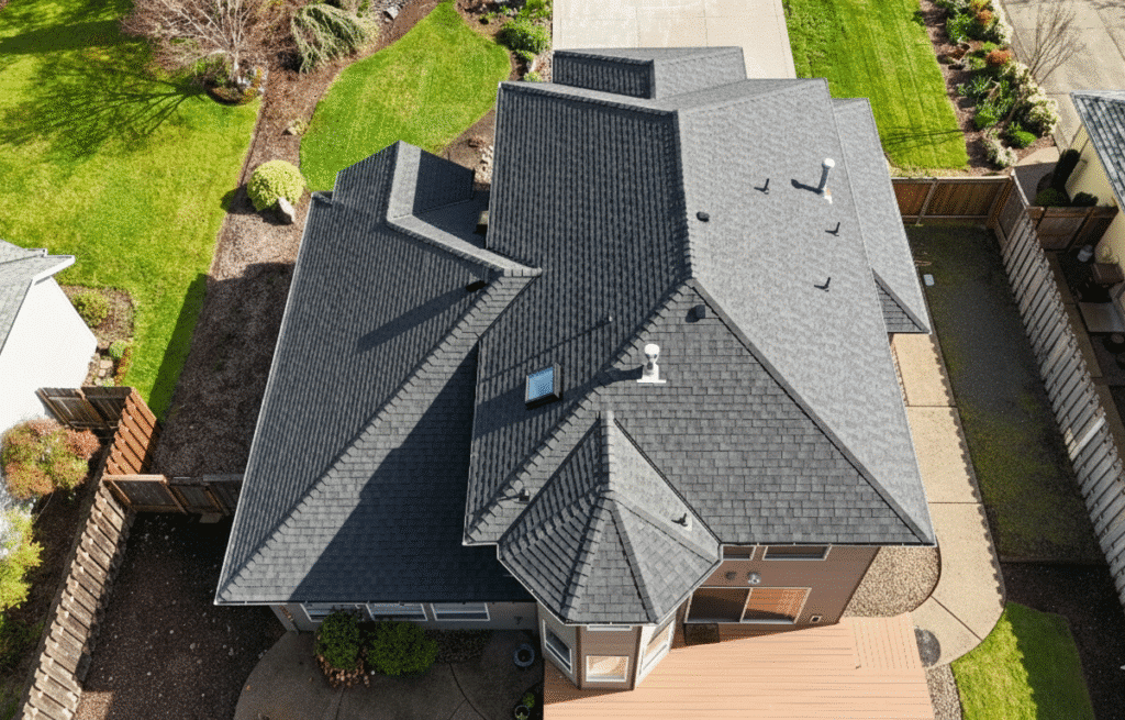 Aerial view of a modern house with a dark gray shingle roof, surrounded by a neatly landscaped yard, wooden fences, a paved driveway, and green lawns.